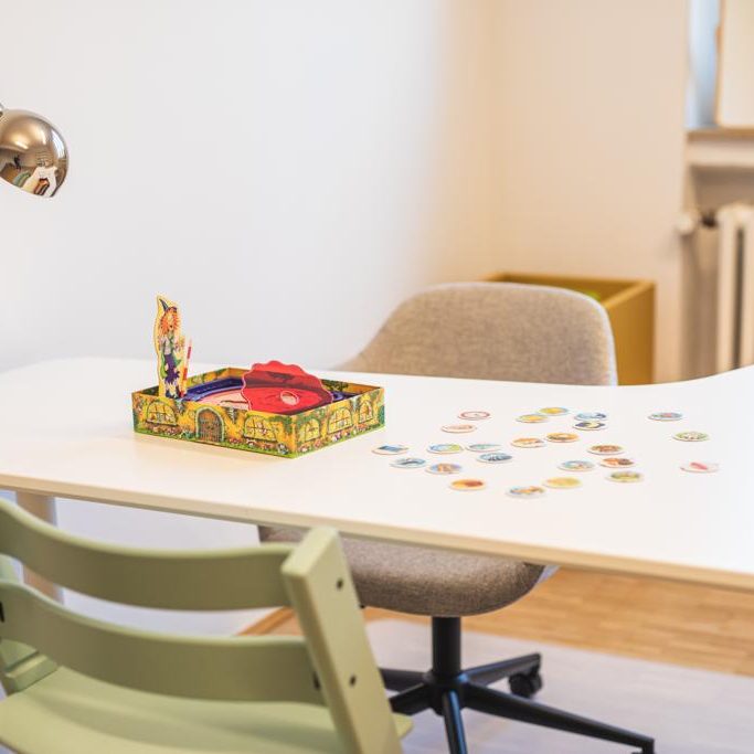 Ein Tisch mit einem Spiel, bunten Spielsteinen und einem Stuhl in einem hellen Raum.